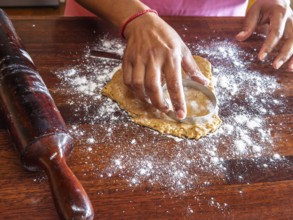Hands using a circular cutter to shape cookie dough on a floured wooden surface. A rolling pin is