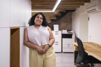 A young woman stands in a modern coworking space with a warm smile. The open office setting is