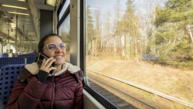 A woman in a cozy jacket is talking on her phone while traveling on a train. She gazes happily out