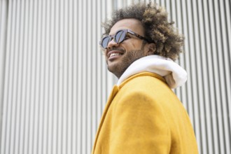 A confident afro man wearing sunglasses and a stylish yellow coat poses in front of a modern