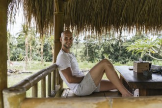 A man in casual attire smiles while relaxing in a bamboo hut, surrounded by lush greenery in Bali.