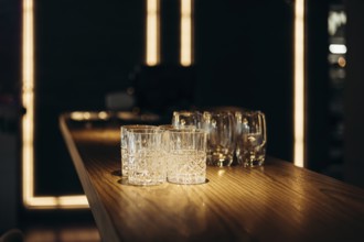 A collection of empty glasses sits on a polished wooden bar, illuminated by warm ambient lighting,