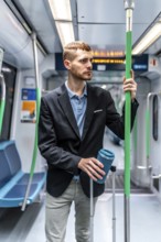 Young businessman commuting on a subway train, holding a reusable coffee cup and luggage, looking