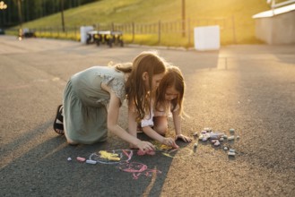 Two kids draw with colorful chalk on asphalt during a bright, sunny day, surrounded by lush