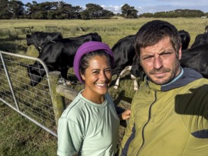 A man and a woman takes a selfie on a sunny day in a grassy field, surrounded by black cows in