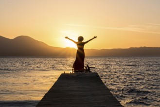 A woman stands on a wooden pier at Papikinou Beach, Milos Island, Greece, embracing the sun as it