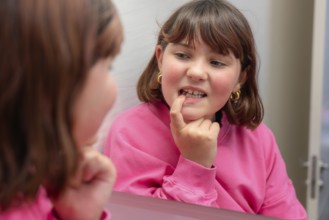 A young girl in a pink sweater smiles as she inspects a loose milk tooth, using a mirror to get a