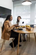Two businesswomen are enjoying breakfast and working together on a laptop in their hotel room