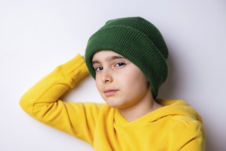 A young child wearing a green knit beanie and a bright yellow hoodie leans against a white wall.