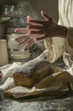 A warm, rustic scene of baking bread. Hands clap to dust flour while a loaf of bread cools on a