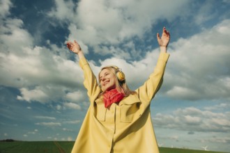 A joyful woman in a yellow coat and red scarf listens to music through headphones, raising her arms