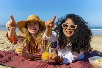 Two cheerful young women lying on a beach blanket, pointing into the distance with excitement while
