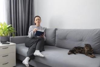 A woman reads a book on a gray couch in a cozy living room with a relaxed cat nearby. The setting