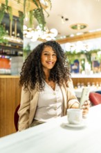 Vertical photo of a happy latin woman using phone while drinking coffee in a modern cafeteria
