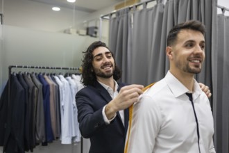 A tailor measures a smiling client suit in a modern suit store. The fitting room features various