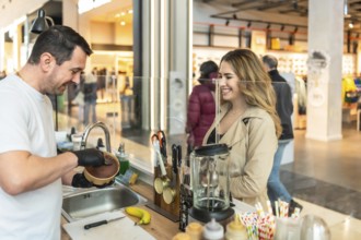 Man in apron serves a bowl of ice cream to a smiling young woman at a mall food stand counter,