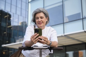 A mature woman with gray hair smiles as she uses a smartphone while standing with an electric