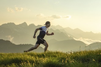 Man in athletic gear running on a grass trail with scenic mountain backdrop. The sun sets behind