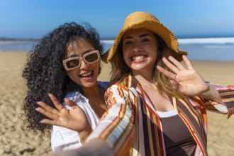 Two cheerful young women on a sunny beach vacation take a joyful selfie, smiling and waving at the