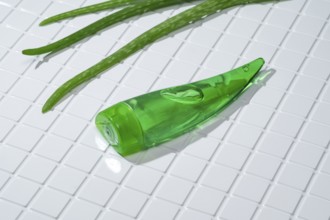 A green aloe vera gel bottle lies alongside fresh aloe leaves on a clean, white-tiled bathroom
