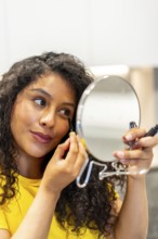 Vertical close-up of a beauty latin young woman making up holding a hand mirror at home