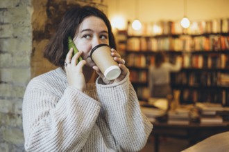 A young woman in a cozy sweater drinks coffee while talking on her smartphone inside a warm