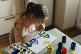 A young girl wearing a pink outfit sits at a clear table, focused intently as she paints a colorful