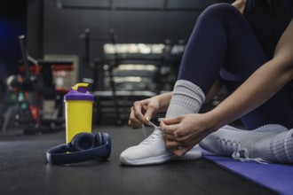A woman ties their white sneakers, preparing for a workout session. Nearby are wireless headphones