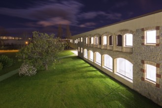 Night view of a well lit hospital with stone and brick facade. The scene includes a green lawn, a