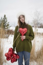 Woman in a green sweater, holding red heart shaped balloons in a snowy park, surrounded by dry