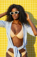 Stylish model wearing a beige bikini and blue striped overshirt, posing against a vibrant yellow