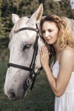 A young woman in a white dress gently embraces a serene white horse outdoors, showcasing a peaceful