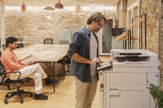 Two professionals in a cozy coworking space, one seated with documents and the other using a copier