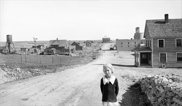 Portrait of Young Girl, Street Scene with Abandoned Grain Mill and Bank (both right) in Background, Mills, New Mexico, USA, Dorothea Lange, Farm Security Administration, May 1935