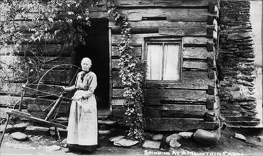 Woman with Spinning Wheel Beside Log Cabin, circa 1910