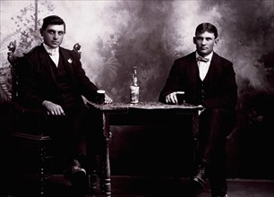 Two Men in Formal Attire Sitting at Table Drinking Beer, circa 1900