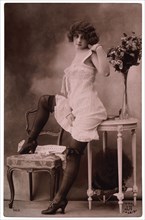 French Lingerie Model Leaning on Table With One Leg Raised on Chair, 1920