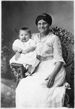 Smiling Woman Wearing Eyeglasses with Happy Infant on Arm of Wicker Chair, Portrait, circa 1920
