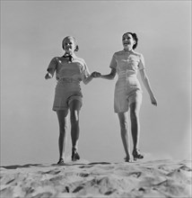 Two Fashionable Women in Shorts Holding Hands While Walking on Beach, circa 1952