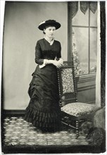 Portrait of Adult Woman in Hat and Long Dress Leaning on Chair Near Window, Tintype, circa 1870