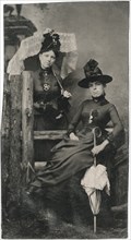 Portrait of Two Adult Women, One Seated, One Standing, in Hats and Long Dresses and Holding Parasols, circa 1870