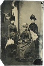 Portrait of Three Adult Women in Hats and Long Dresses with Parasols, one Seated, circa 1870