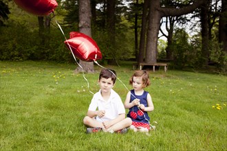 Young Boy and Girl Sitting on Grass and Holding Red Balloons