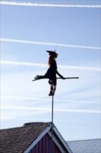 Witch and Broom on Pole Attached to Barn Roof with Airplane Trails in Sky