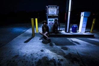 Young Adult Woman in Hat Sitting Near Gas Pump at Gas Station at Night