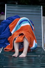 Young Boy Hiding Under Striped Towel on Pool Chair