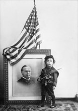 Boy in Sailor Uniform holding Sword in front of flag-draped, head-and-shoulders portrait of William McKinley, 1896