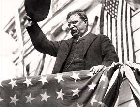 Theodore Roosevelt on a flag-draped platform, waving his hat during his tour through New Jersey before the Progressive Party Convention, September 1912