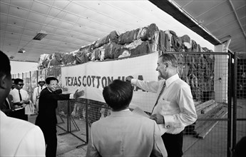 U.S. Vice President Lyndon Johnson Visiting Textile Mill with Vietnamese men standing in front of a large quantity of textiles with a "Texas Cotton U.S.A." Banner, Saigon, South Vietnam, photograph by...