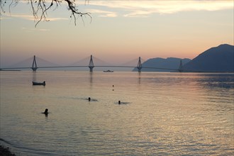 Silhouette of Three People Swimming with Rio–Antirrio Bridge in Background at Sunset, Patras, Greece